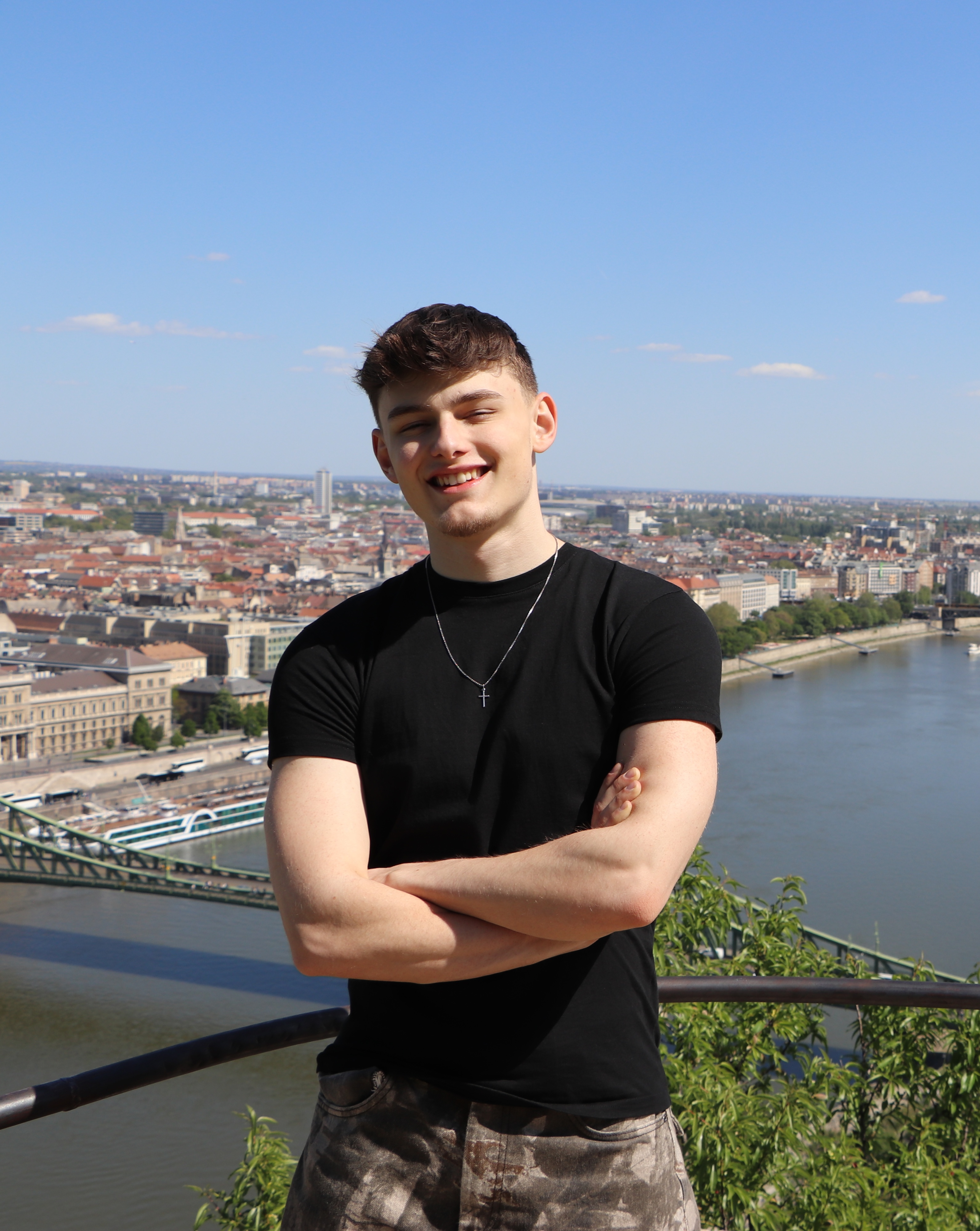 A smiling young man in a black t-shirt and camo shorts with arms crossed, overlooking Budapest and the Danube.